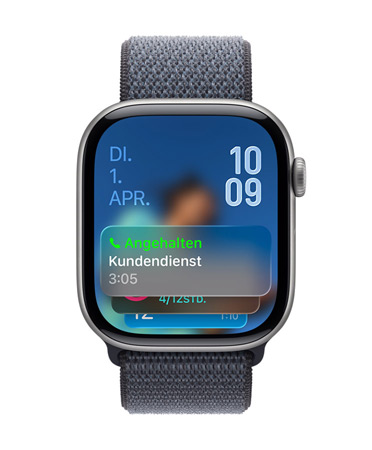 Eine Apple Watch in Space Grau mit Armband in Grau, das Zifferblatt zeigt unten die Benachrichtigung für die Warteschleifenassistenz, das Datum ist oben links, Zeitziffern sind gestapelt oben rechts zu sehen