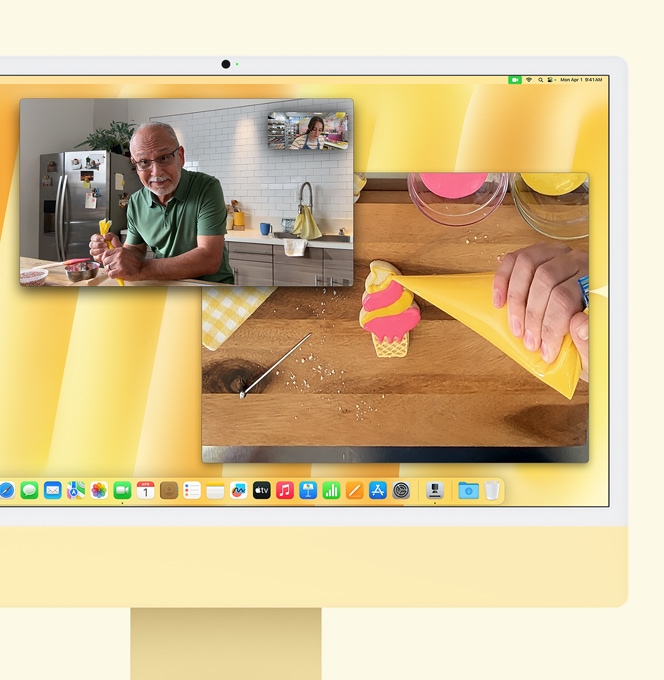 Żółty iMac. Demonstracja funkcji Widok blatu. Na ekranie widoczna jest piekarka podczas rozmowy wideo z inną osobą. W jednym oknie widać piekarkę i twarz mężczyzny. W drugim oknie widoczne są ręce piekarki lukrującej ciastko.