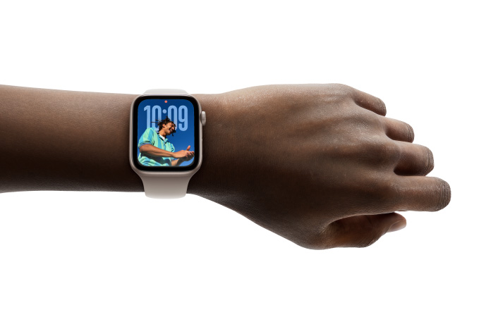 ภาพระยะใกล้ของ Apple Watch สีเงินพร้อมสายที่ทำจากยางบนข้อมือของบุคคล, แสดงแอปเวลาในดีไซน์แบบ Liquid Glass ใหม่