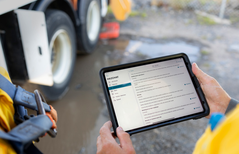 Hænder holder en iPad med HazChat-appen åben, hvor der vises joboplysninger, planlægning, risici og foranstaltninger, mens en person står på en byggeplads ved en mudret vandpyt